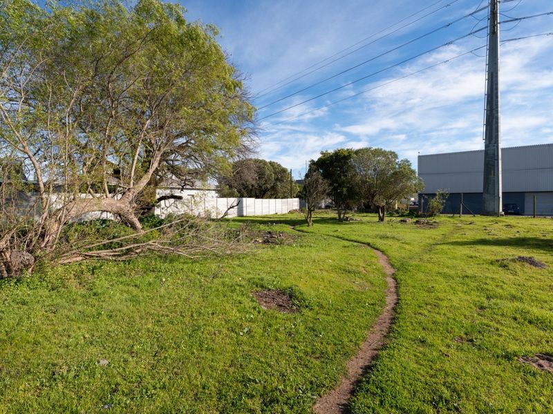 A vacant residential plot featuring grassy terrain, mature trees, and perimeter walling, situated near industrial infrastructure.