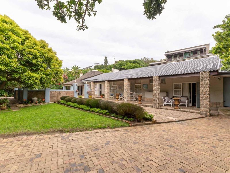 Rear view of the property featuring a covered patio with stone pillars, a paved driveway, and a well-maintained garden area.