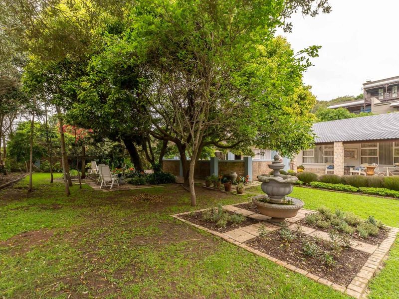 A well-maintained garden featuring a central stone fountain, mature trees, manicured lawn, and a stone-clad patio area attached to the main residence.