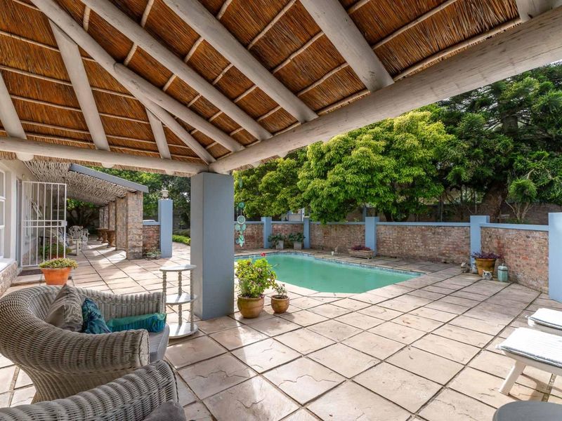 Covered patio area with a thatched roof overlooking a swimming pool and private garden, featuring paved flooring and brick boundary walls.