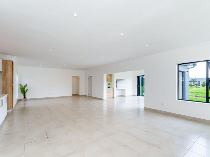 Spacious open-plan living area featuring neutral tiled flooring, recessed lighting, and a modern kitchen with built-in cabinetry, leading to an indoor braai area.