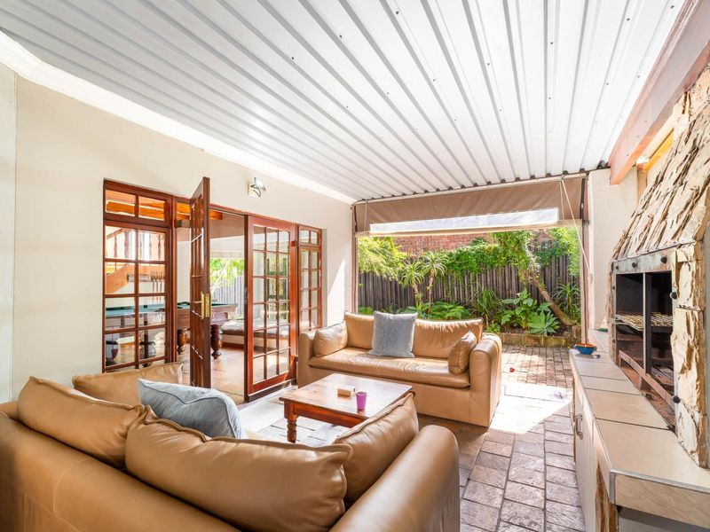 Covered outdoor patio featuring a built-in braai with stone cladding, tiled flooring, and timber-framed glass doors leading to the interior.