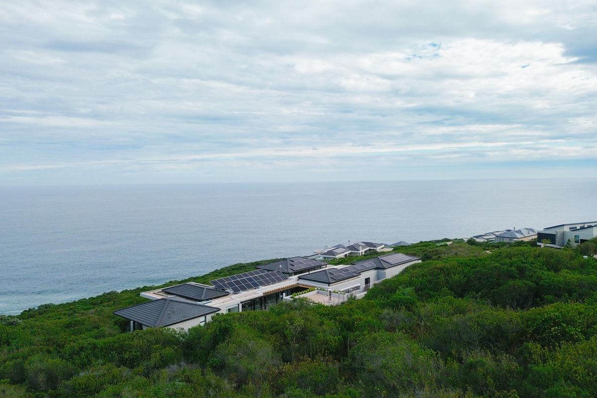 Aerial view of a modern coastal residence featuring extensive solar panel installations on the roof, set within a lush natural landscape overlooking the ocean.