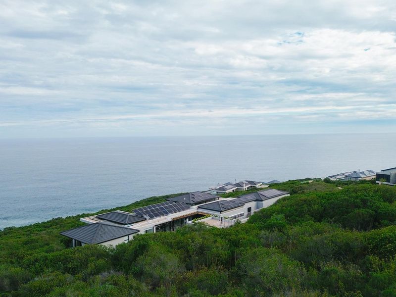 Aerial view of a modern coastal residence featuring extensive solar panel installations on the roof, set within a lush natural landscape overlooking the ocean.
