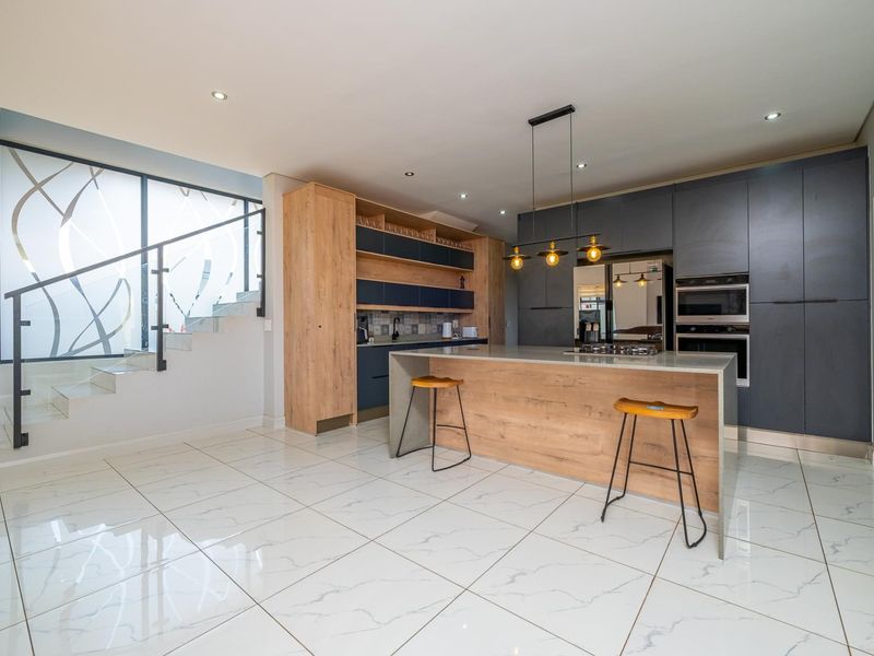Modern open-plan kitchen featuring a central island, dark cabinetry, integrated appliances, and sleek marble-look tiled flooring, adjacent to a contemporary staircase.