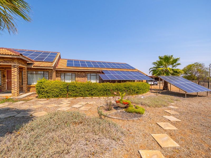 Rear view of a brick farmhouse featuring extensive roof-mounted and ground-mounted solar panel arrays, a paved garden path, and established landscaping.