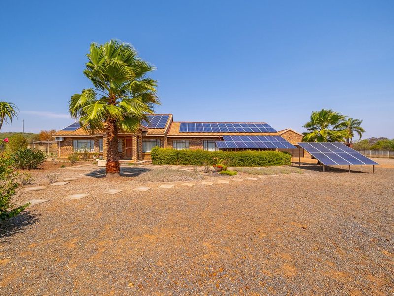 Front view of a brick farmhouse featuring extensive roof-mounted and ground-mounted solar panel arrays, set within a landscaped garden with mature palm trees.