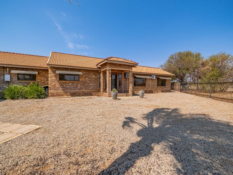 A well-maintained single-story face brick farmhouse featuring a tiled roof, covered entrance, and a spacious gravel driveway enclosed by metal fencing.