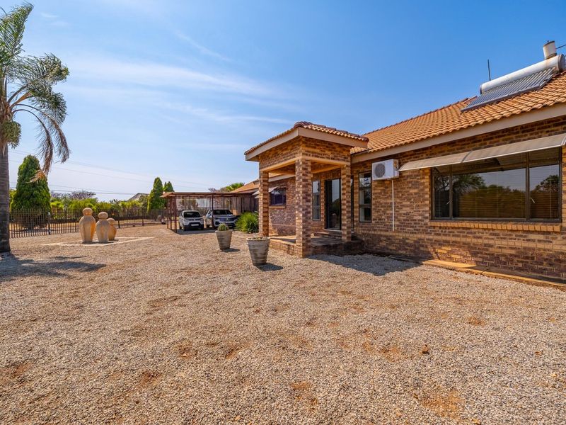Rear view of a face brick farm house featuring a covered patio, solar water heater, and a spacious gravel yard with an attached carport.