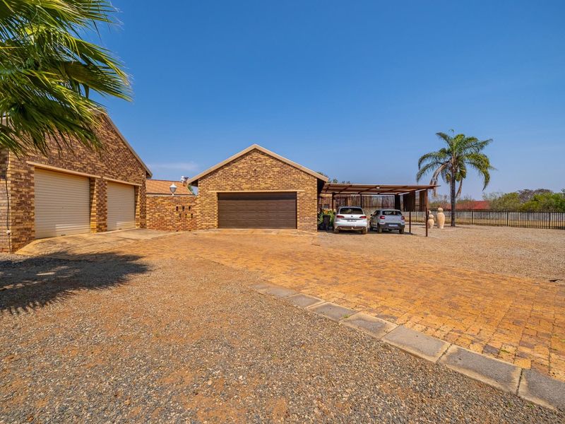 Spacious farm property exterior featuring a brick facade, multiple garage bays, a covered carport, and an expansive paved and gravel driveway area.