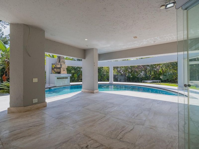 Spacious covered patio area featuring stone tile flooring and a built-in outdoor fireplace, overlooking a curved swimming pool and landscaped garden.