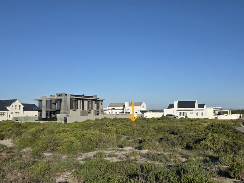 A vacant residential plot situated in a coastal estate, featuring natural vegetation and adjacent to a wooden boardwalk with surrounding modern homes.