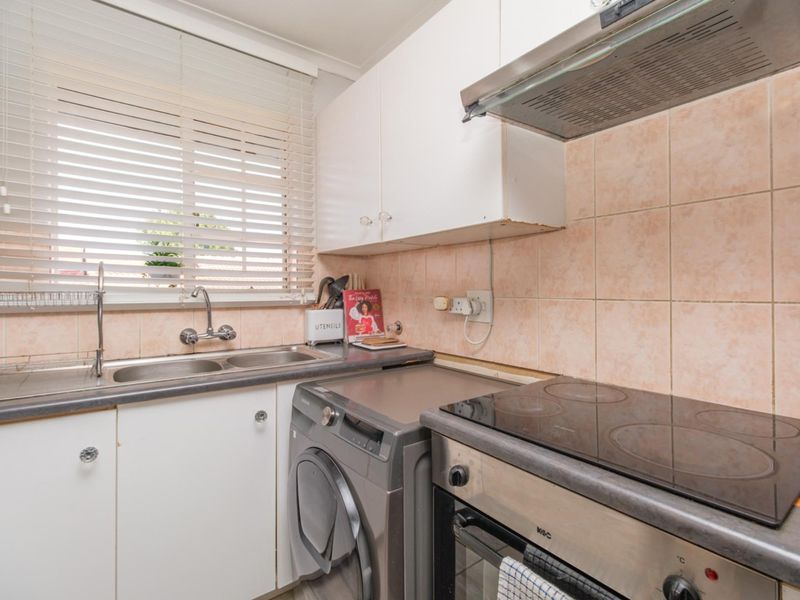 Compact kitchen featuring built-in cabinetry, electric hob, oven, extractor fan, and a double sink with tiled backsplash and venetian blinds.