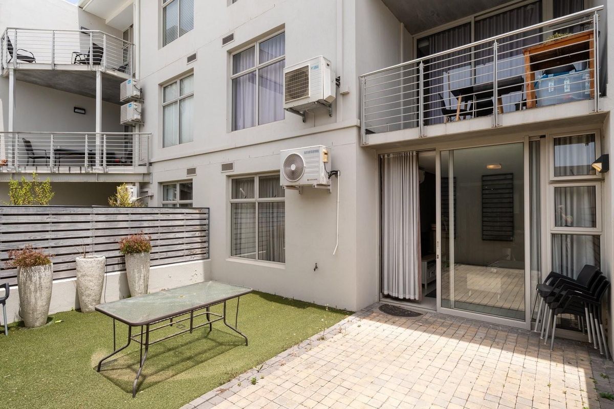 Rear exterior view of an apartment featuring a paved patio, artificial lawn, timber fencing, and access to the living area via sliding glass doors.