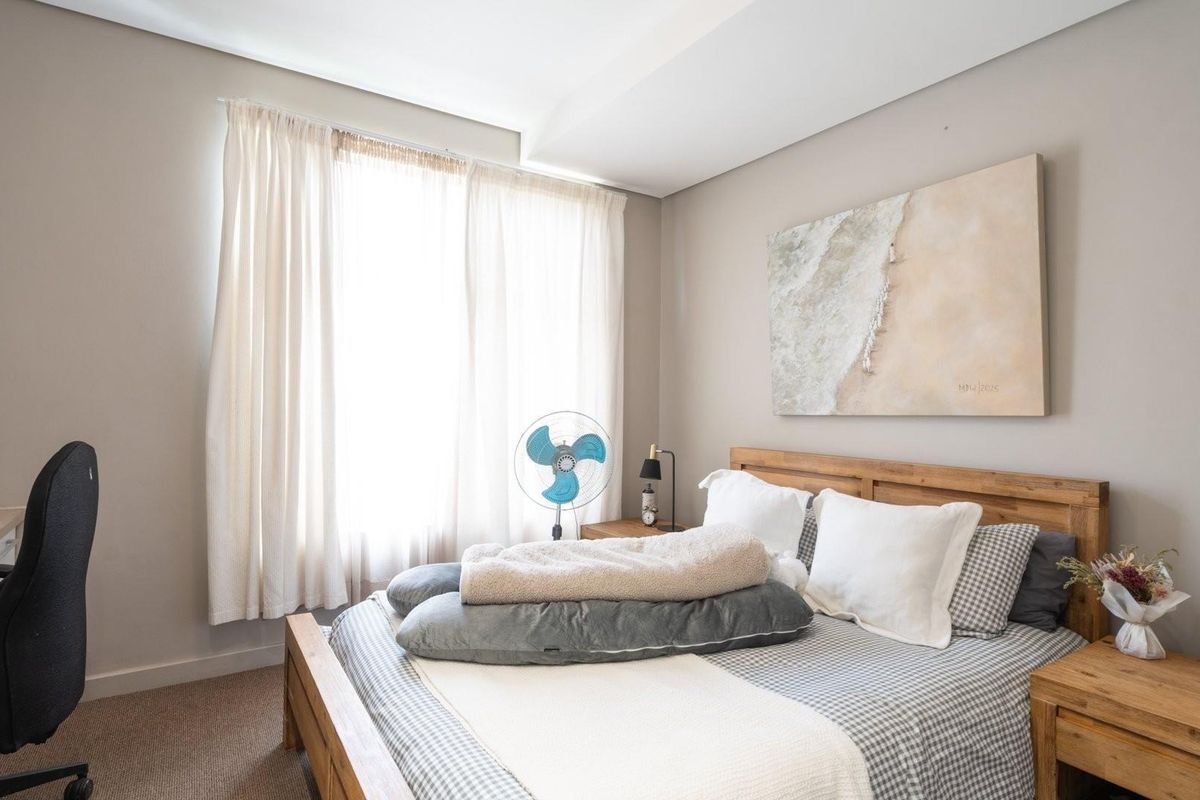 A well-lit bedroom featuring neutral wall tones, carpeted flooring, and large windows with curtains, providing a comfortable and functional living space.