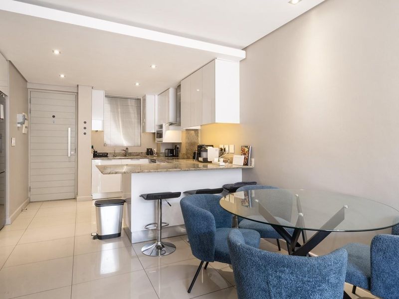Modern open-plan kitchen and dining area featuring glossy white cabinetry, granite countertops, and sleek porcelain floor tiles in a well-lit, contemporary apartment setting.