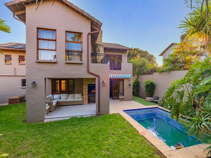 Rear view of a double-story house featuring a covered patio with built-in braai, a swimming pool, and a well-maintained garden.