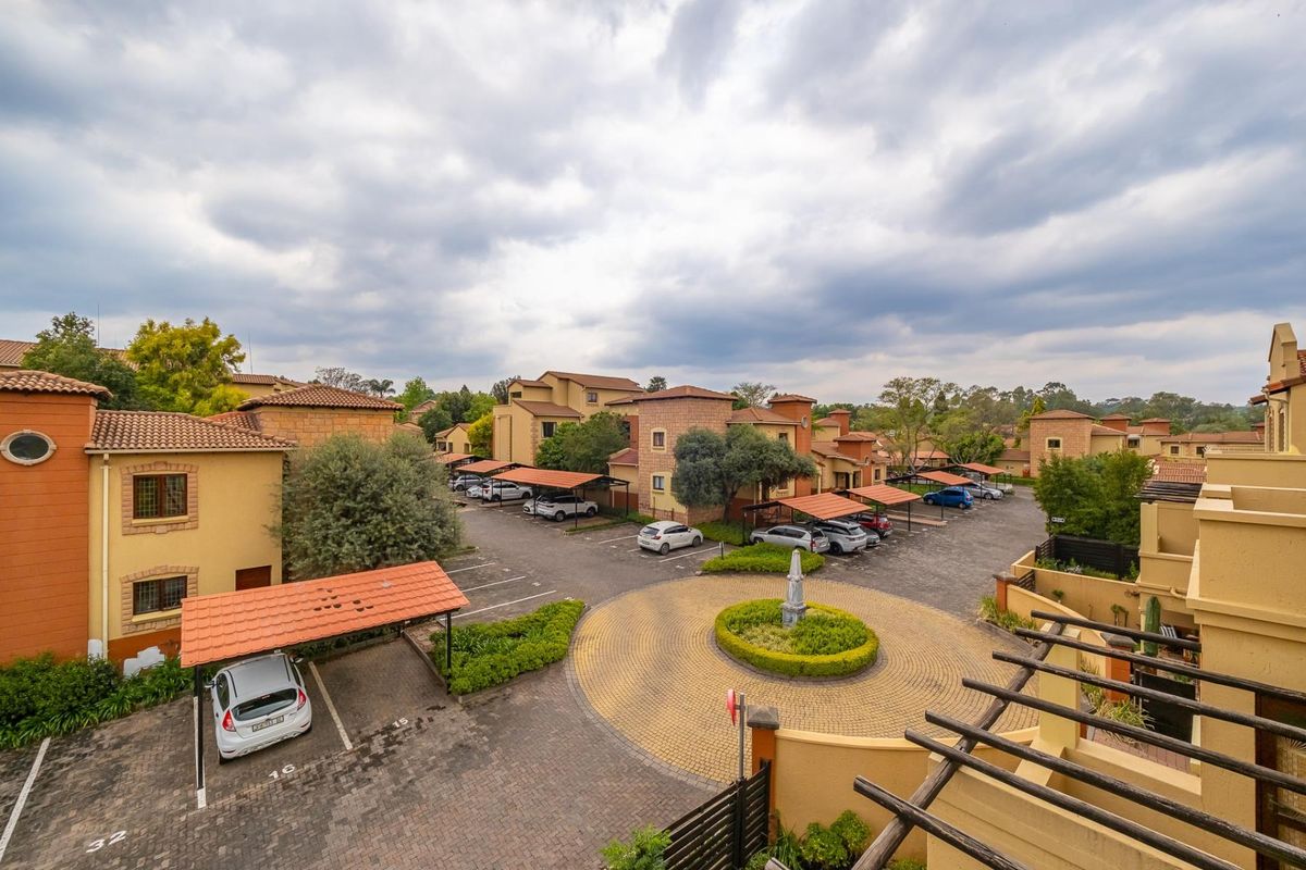 An elevated aerial view of the apartment complex grounds, featuring paved parking areas, covered carports, and a central landscaped roundabout.