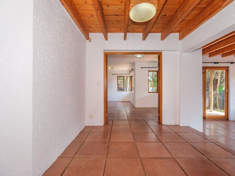 A bright, empty hallway featuring exposed timber ceiling beams and terracotta floor tiles, leading into adjacent living spaces with timber-framed windows.