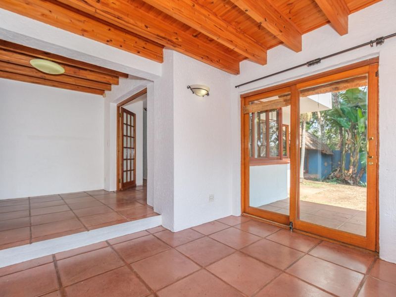 A bright, empty patio area featuring exposed timber beam ceilings, terracotta floor tiles, and timber-framed glass doors leading to the garden.