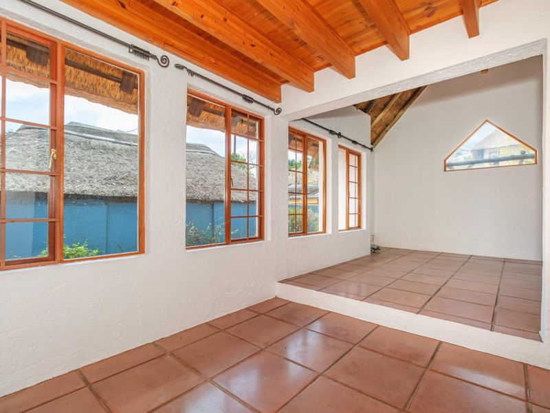 A bright, empty living area featuring exposed timber ceiling beams, terracotta floor tiles, and large timber-framed windows with a unique triangular feature window.