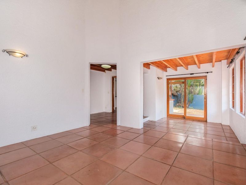 An empty living area featuring terracotta floor tiles, exposed timber ceiling beams, and a sliding glass door providing access to the garden.