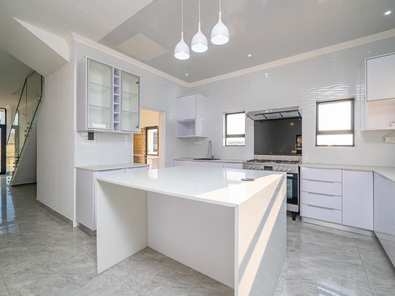 Modern kitchen featuring a central quartz island, sleek white cabinetry, built-in gas hob, and high-quality tiled flooring, adjacent to a glass-railed staircase.