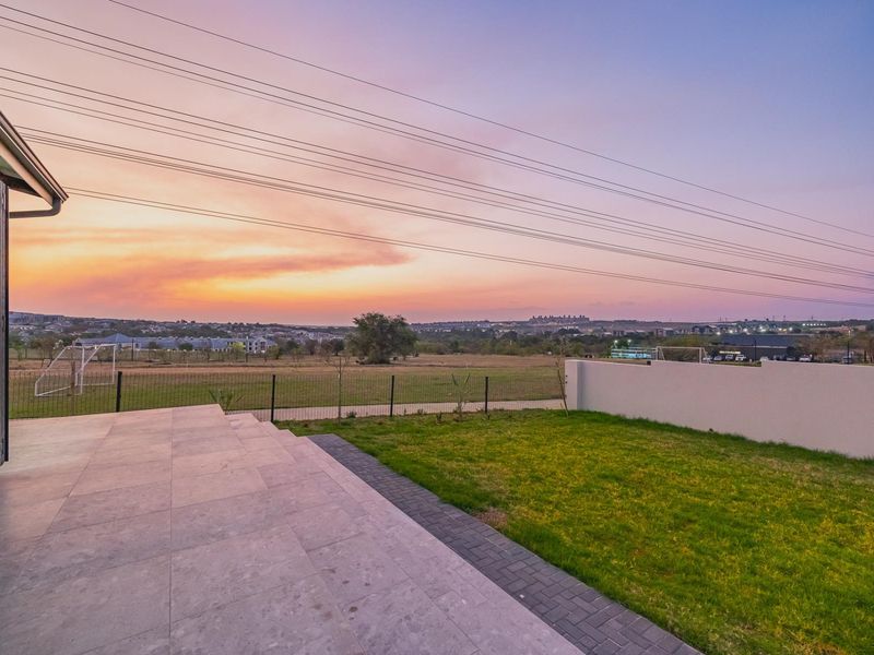 A rear exterior view featuring a tiled patio, a small lawn, and a boundary wall overlooking an open landscape at sunset.