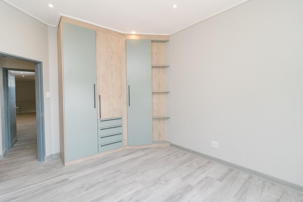 A bright, modern bedroom featuring light wood-tone laminate flooring, neutral walls, and a contemporary built-in wardrobe with integrated shelving and drawers.
