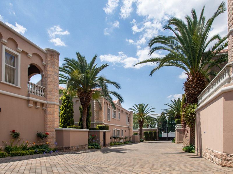 A well-maintained paved driveway leading through an apartment complex, featuring stone-clad pillars, decorative balustrades, and established palm trees.