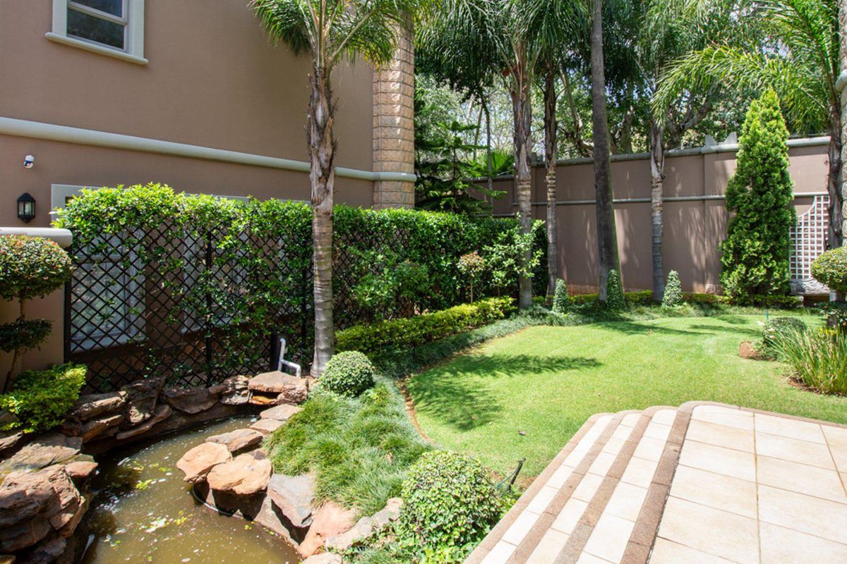 A well-maintained rear garden featuring a stone water feature, manicured lawn, tiled patio area, and established palm trees enclosed by a perimeter wall.