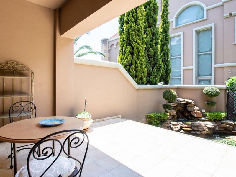 A covered patio area featuring tiled flooring and a built-in stone water feature, overlooking a landscaped garden area.
