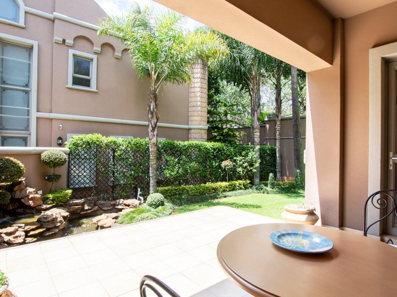 A covered patio area featuring tiled flooring, overlooking a landscaped garden with a built-in water feature and established greenery.