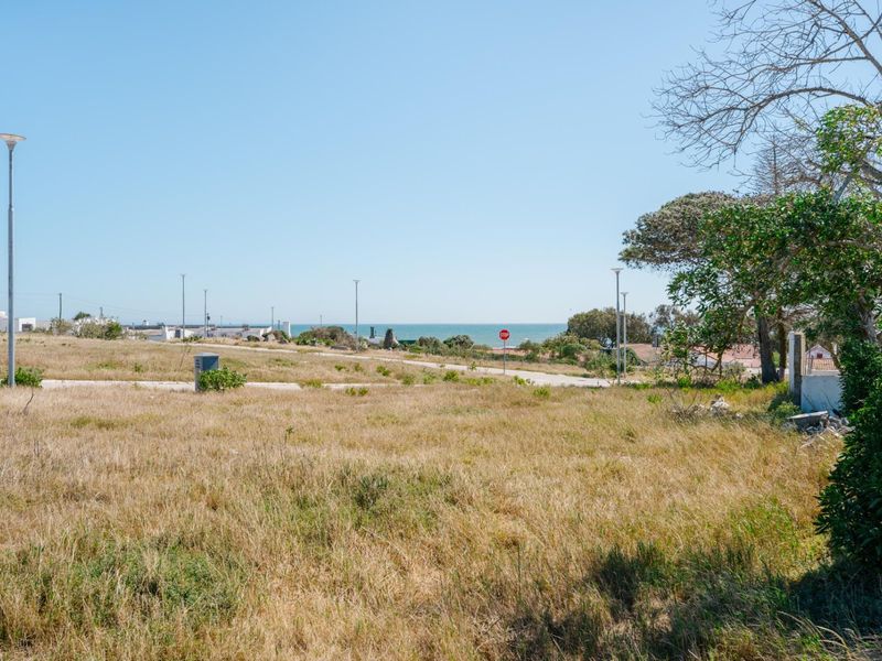 A view of a vacant residential plot featuring dry grass, street lighting, and a distant ocean horizon.