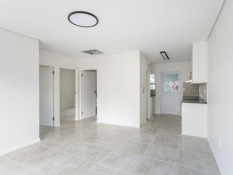 Bright open-plan living area featuring neutral tiled flooring, modern flush-mount lighting, and a compact kitchen with built-in cabinetry and hob.