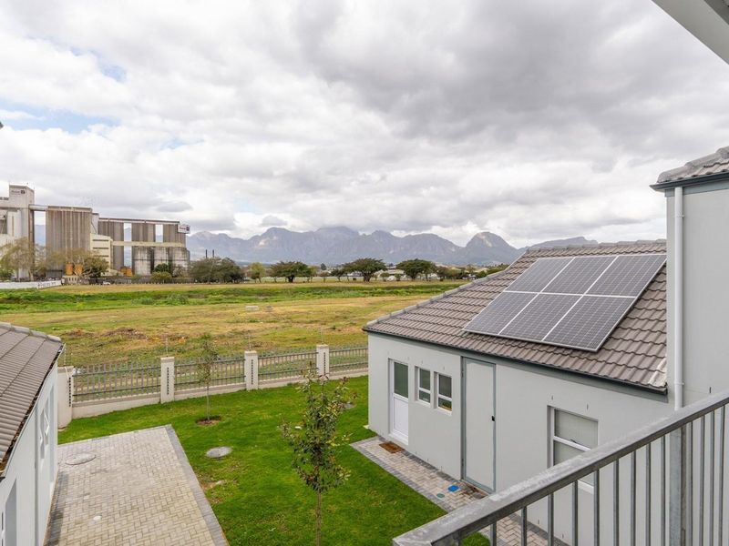 Elevated view from a balcony overlooking a landscaped garden, neighboring property with solar panels, and scenic mountain landscape in the distance.