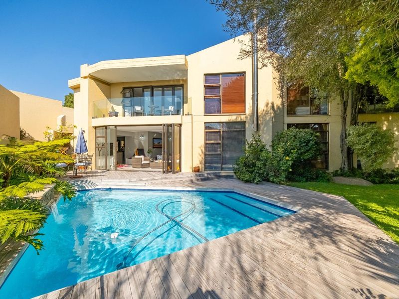 Rear view of a modern two-story house featuring a swimming pool, wooden deck, glass balcony, and large bi-fold doors opening to the living area.