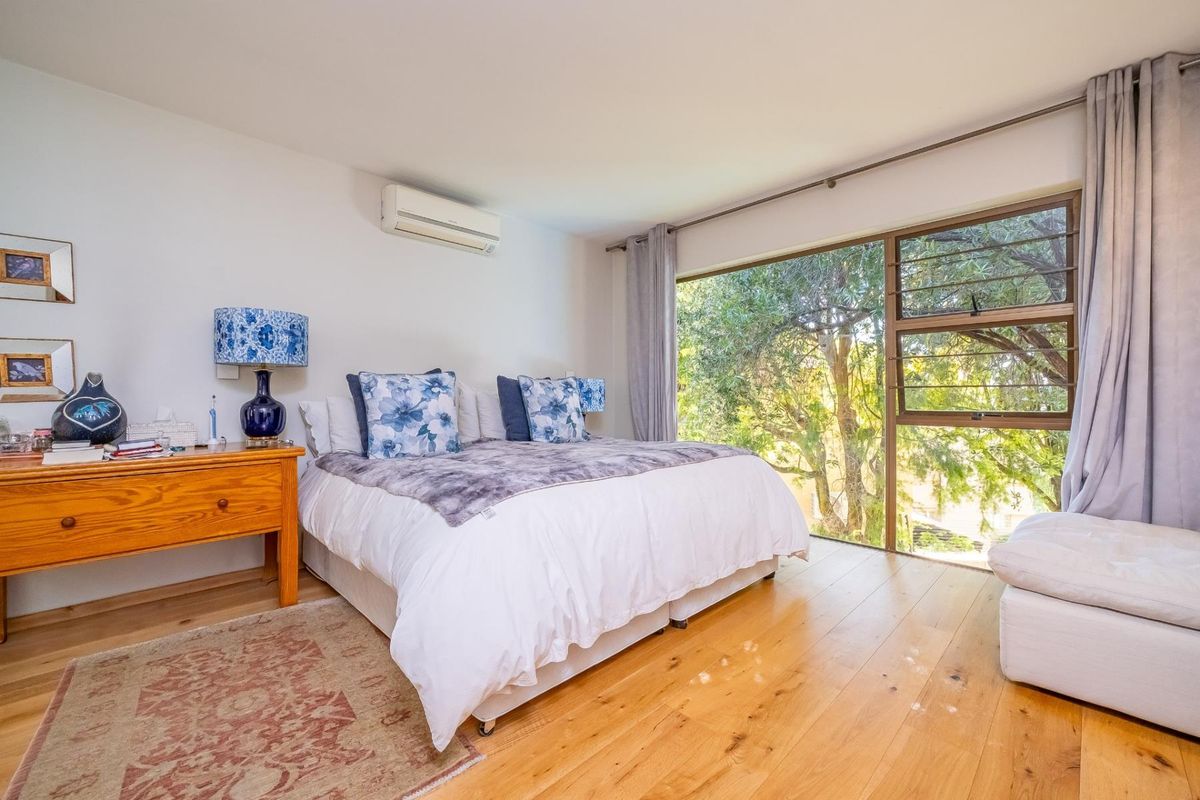 A bright and spacious bedroom featuring polished hardwood flooring, a wall-mounted air conditioning unit, and a large window offering pleasant views of mature trees.