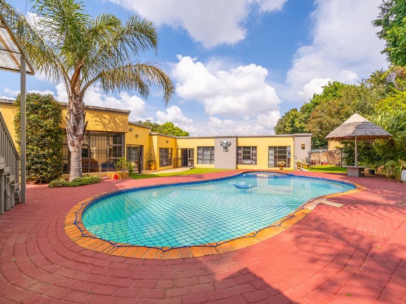 A spacious outdoor pool area featuring a swimming pool with safety net, red brick paving, and a thatched lapa set against the rear building facade.