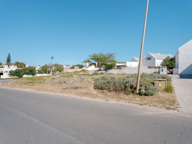 Street view of a vacant residential plot featuring a paved driveway entrance, boundary wall, and utility connections in a established neighborhood.