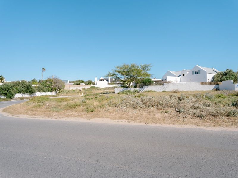 A street-level view of a vacant residential plot featuring natural vegetation and surrounding boundary walls in a coastal neighborhood.