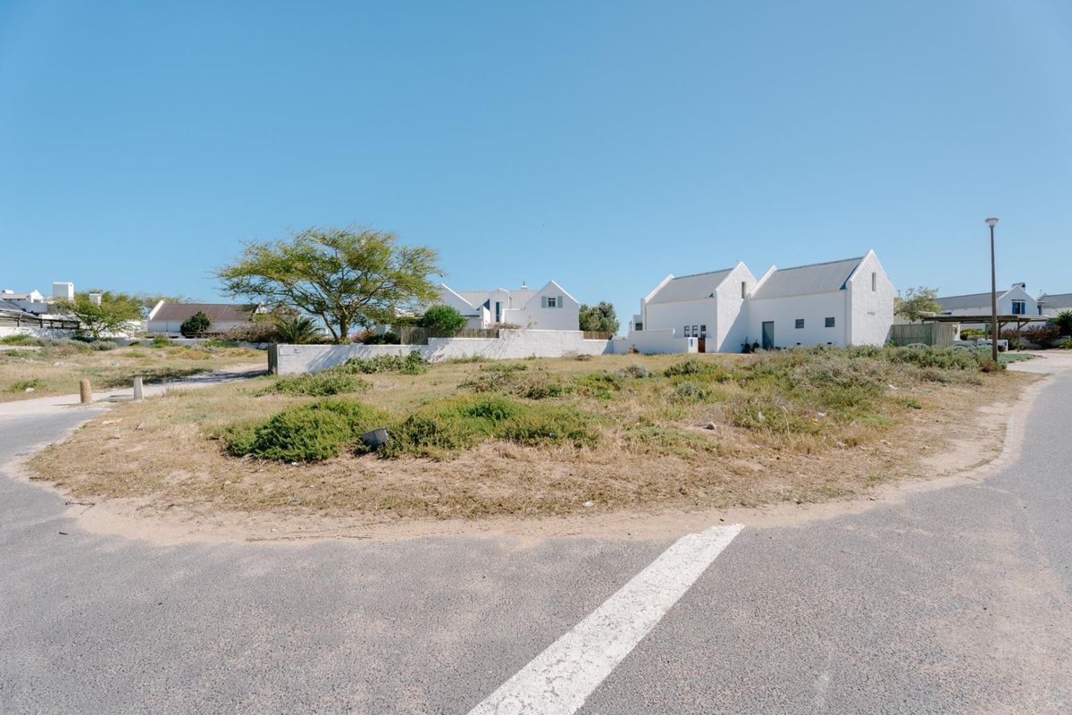 A street-level view of a vacant residential plot featuring natural vegetation, a boundary wall, and adjacent white-walled coastal style homes.