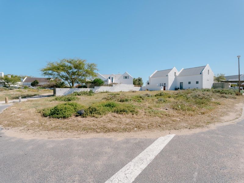 A street-level view of a vacant residential plot featuring natural vegetation, a boundary wall, and adjacent white-walled coastal style homes.