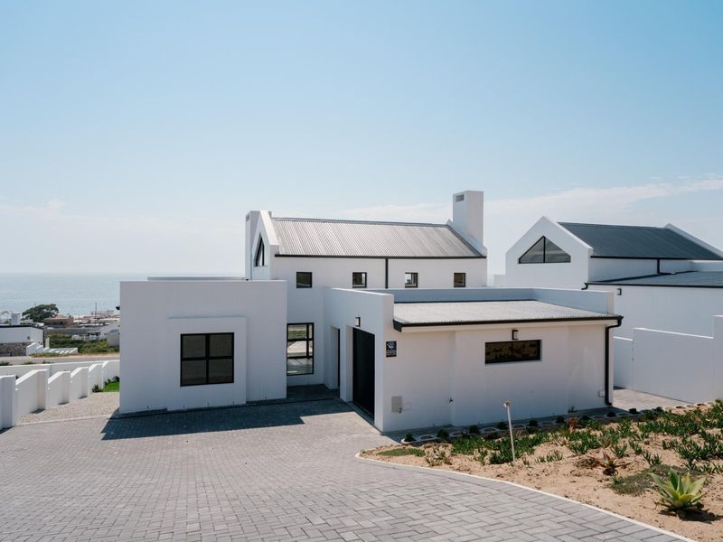 Modern white coastal home featuring a paved driveway, metal roofing, and clean architectural lines with ocean views in the background.