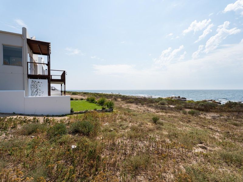A rear view of a coastal property featuring a balcony with a pergola, overlooking a natural dune landscape and the ocean.