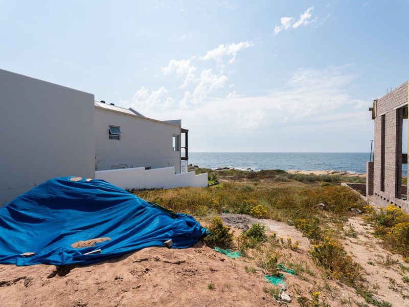 A view of a vacant residential plot featuring coastal scenery, adjacent modern housing, and ongoing construction work on the neighboring property.