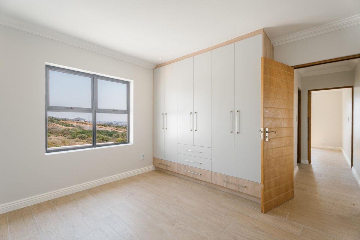 A bright, modern bedroom featuring neutral tiled flooring, built-in cabinetry with a light wood finish, and a large window offering scenic views.