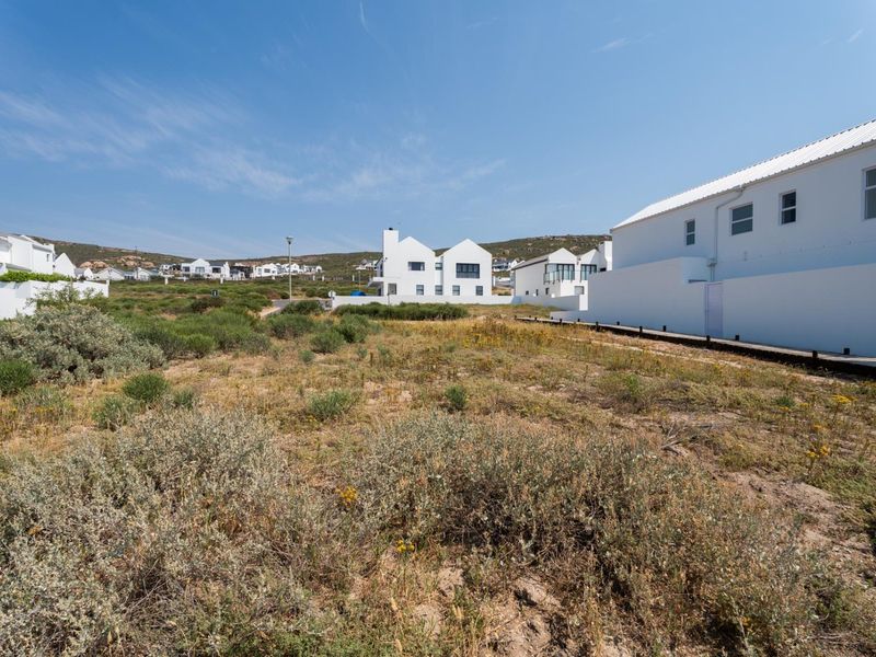 A view of a vacant residential plot featuring natural vegetation, a wooden walkway, and surrounding modern white residential architecture.