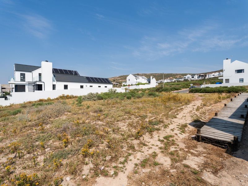 A vacant residential plot featuring a wooden boardwalk path, surrounded by established coastal homes with solar installations in a quiet neighborhood setting.