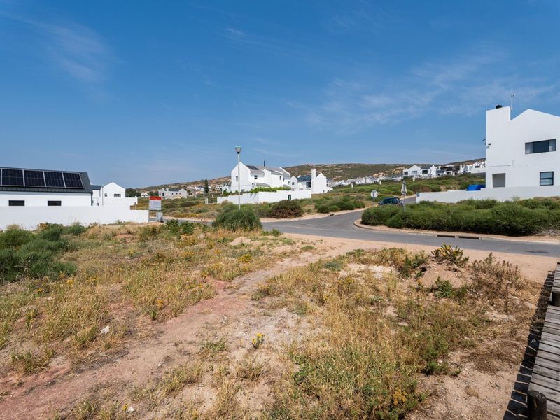 A view of a vacant residential plot featuring a wooden boardwalk, adjacent to a paved street in a coastal estate setting.